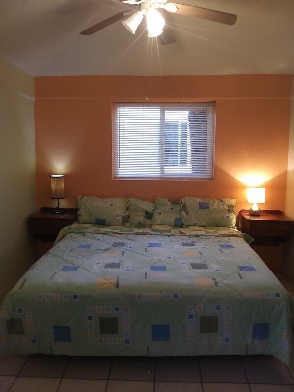 Une chambre à coucher comprenant un lit, deux tables de chevet, un ventilateur de plafond, une fenêtre avec des stores et une lampe murale.