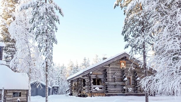 Lumihuntuun peittynyt tukkimökki, jonka edessä on kyltti, jossa lukee ”TEERITAIVAS”.