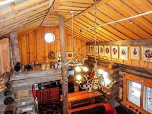 Acogedor interior de cabaña de troncos con techo de madera, una mesa de comedor, sillas, una araña y cuadros en las paredes.