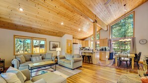 Amplia sala de estar con techo de madera, cocina y ventanas grandes con vistas al exterior.