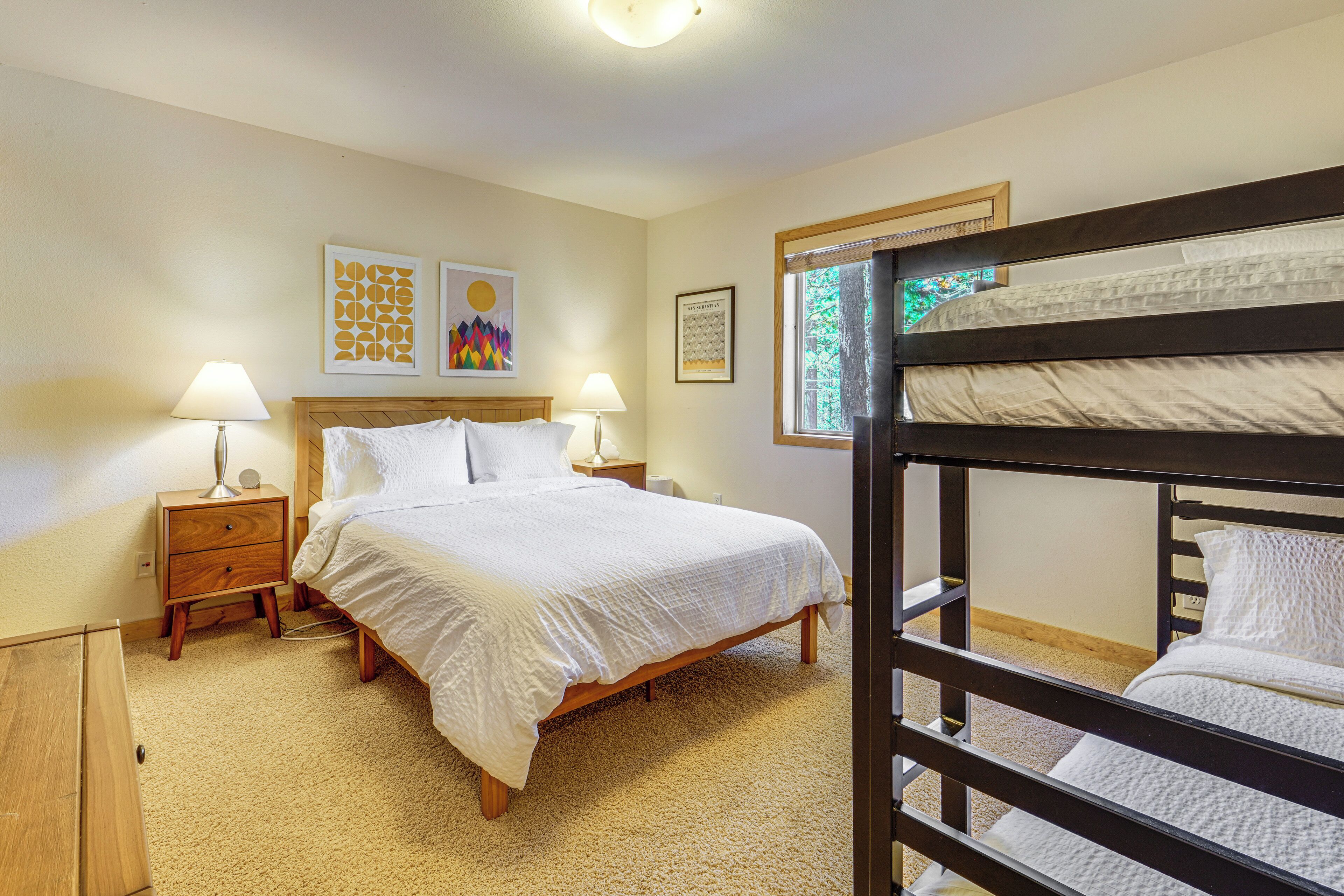 A bedroom with a bunk bed, a single bed, two bedside tables with lamps, and artwork on the walls.