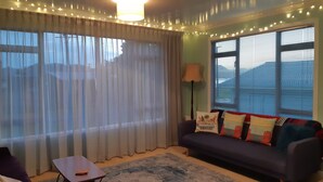 Un salon avec un canapé, une petite table et une lampe. Il y a des rideaux et une fenêtre avec des stores.