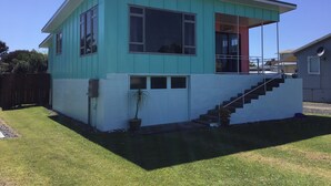 Une maison à deux étages, à la façade turquoise et blanche, avec une porte rouge et un petit jardin devant, où se trouve un pot de fleurs.