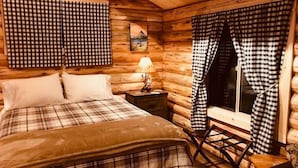 Ein gemütliches Schlafzimmer in einer Blockhütte mit einem Bett, einem Nachttisch, einem Sessel und karierten Vorhängen.