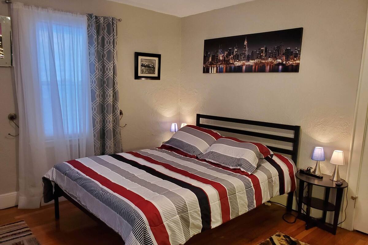 A bedroom with a bed, two bedside lamps, a framed picture, and a cityscape painting on the wall.