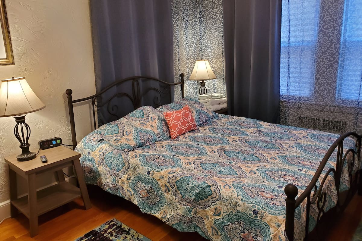 A bedroom with a bed, two bedside lamps, a clock, and a window with curtains.