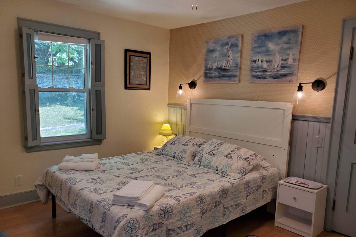 A bedroom with a bed, a nightstand, a lamp, a window, and two paintings on the wall.