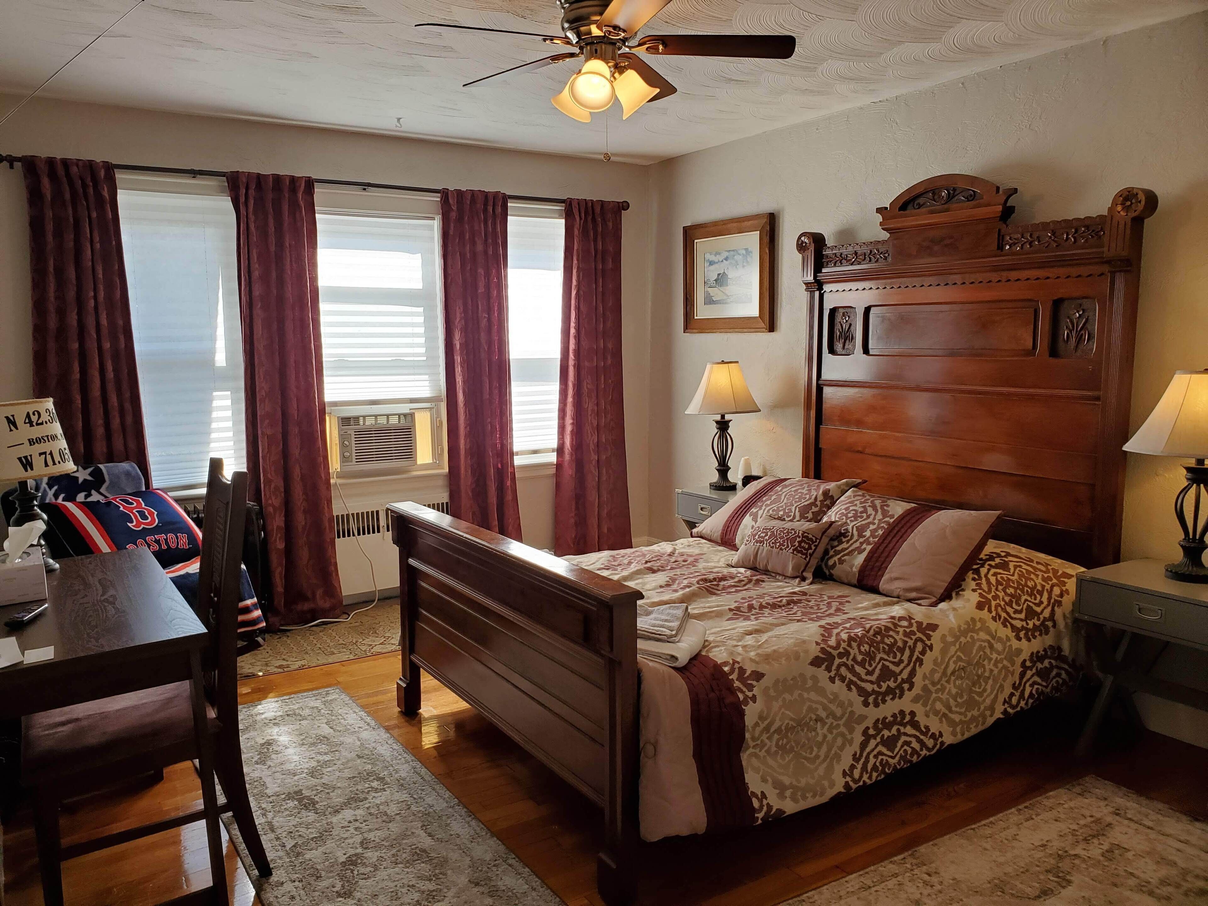 A bedroom with a wooden bed, a desk, a chair, a ceiling fan, and a window with curtains.