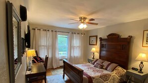 Quarto com cama de madeira, escrivaninha, cadeira, ventoinha no teto e janela com cortinas.