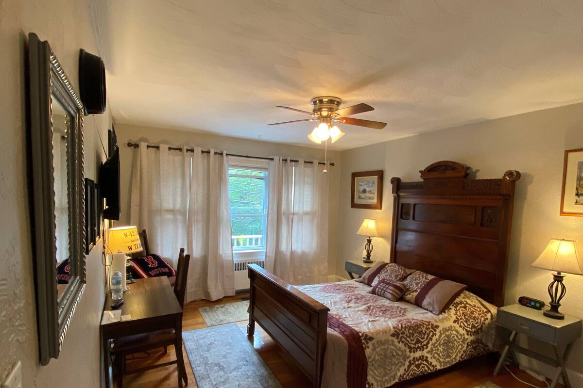 A bedroom with a wooden bed, a desk, a chair, a ceiling fan, and a window with curtains.
