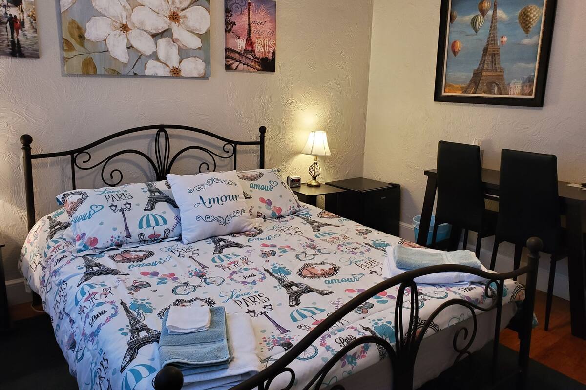 A neatly made bed with Paris-themed bedding, a bedside lamp, a small table, and a chair.