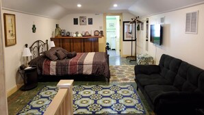 Quarto com cama, sofá preto, mesinha de madeira, tapete estampado e TV fixada na parede.