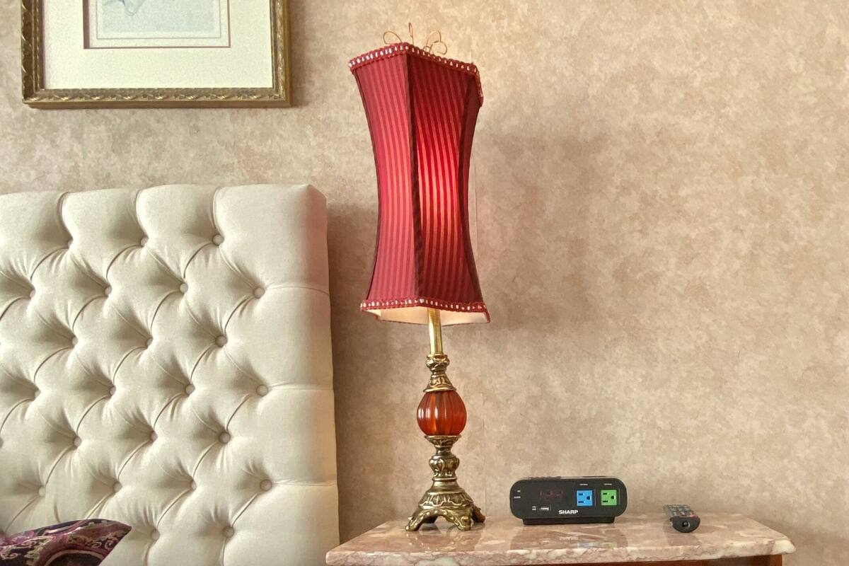 A bedroom with a tufted headboard, a red lamp, a clock, a bedside table with a Sharp power strip, and a patterned bedspread.