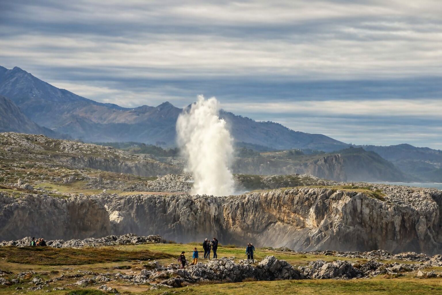 Un géiser con un grupo de personas observándolo en un paisaje montañoso y rocoso.