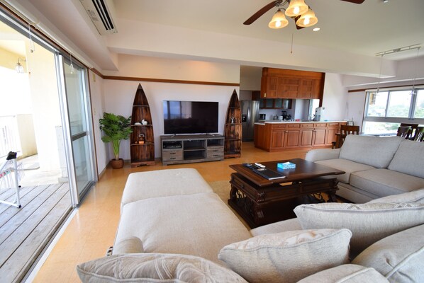 A spacious living room with a large flat-screen TV, a wooden dining table, and a grey sofa.