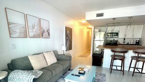 A modern living room with a grey sofa, a blue coffee table, and a kitchen area with a dining table and bar stools. - One block to Vanderbilt Beach! (Naples)