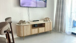 A living room with a flat-screen TV on the wall, a wooden entertainment unit, a dining area with bar stools, and a large window with curtains. - One block to Vanderbilt Beach! (Naples)
