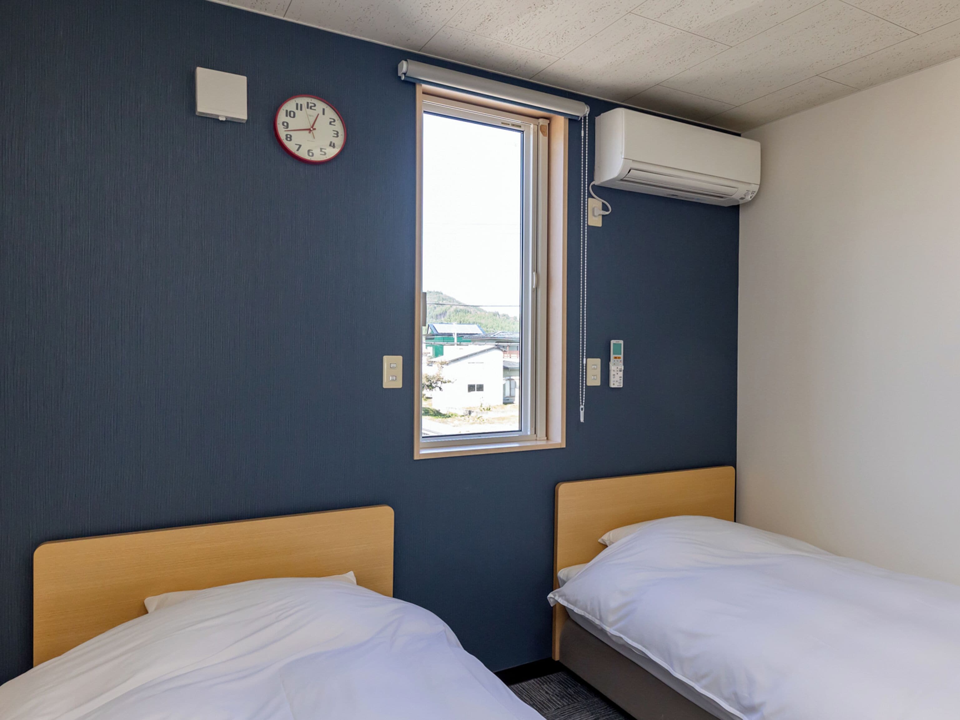 Hotellihuone, jossa on kaksi erillistä sänkyä, herätyskello, ilmastointilaite ja ikkuna, josta on näkymä ulkona oleviin rakennuksiin.