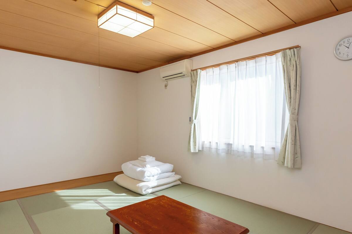 Ein Zimmer mit Tatami-Boden, einem Holztisch, einem Stapel weißer Handtücher, einem Fenster mit durchsichtigen Vorhängen und einer an der Wand befestigten Uhr.