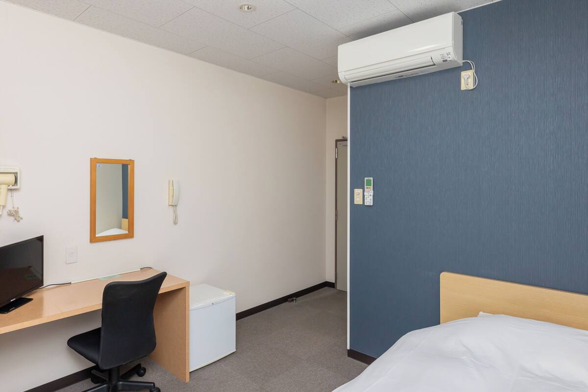 Ein Hotelzimmer mit einem Bett, einem Schreibtisch mit Stuhl, einem kleinen Kühlschrank, einem Spiegel, einem Telefon und einer Klimaanlage.