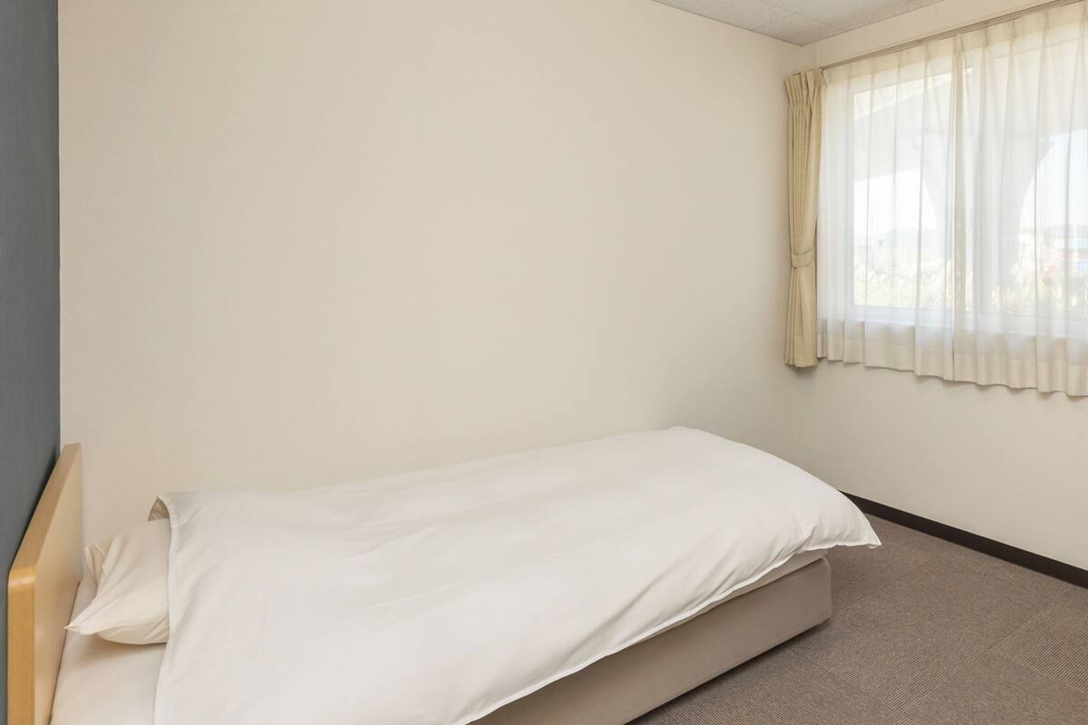 Ein kleines Zimmer mit einem Einzelbett, einer schlichten weißen Wand, einem Fenster mit durchsichtigen Vorhängen und Teppichboden.