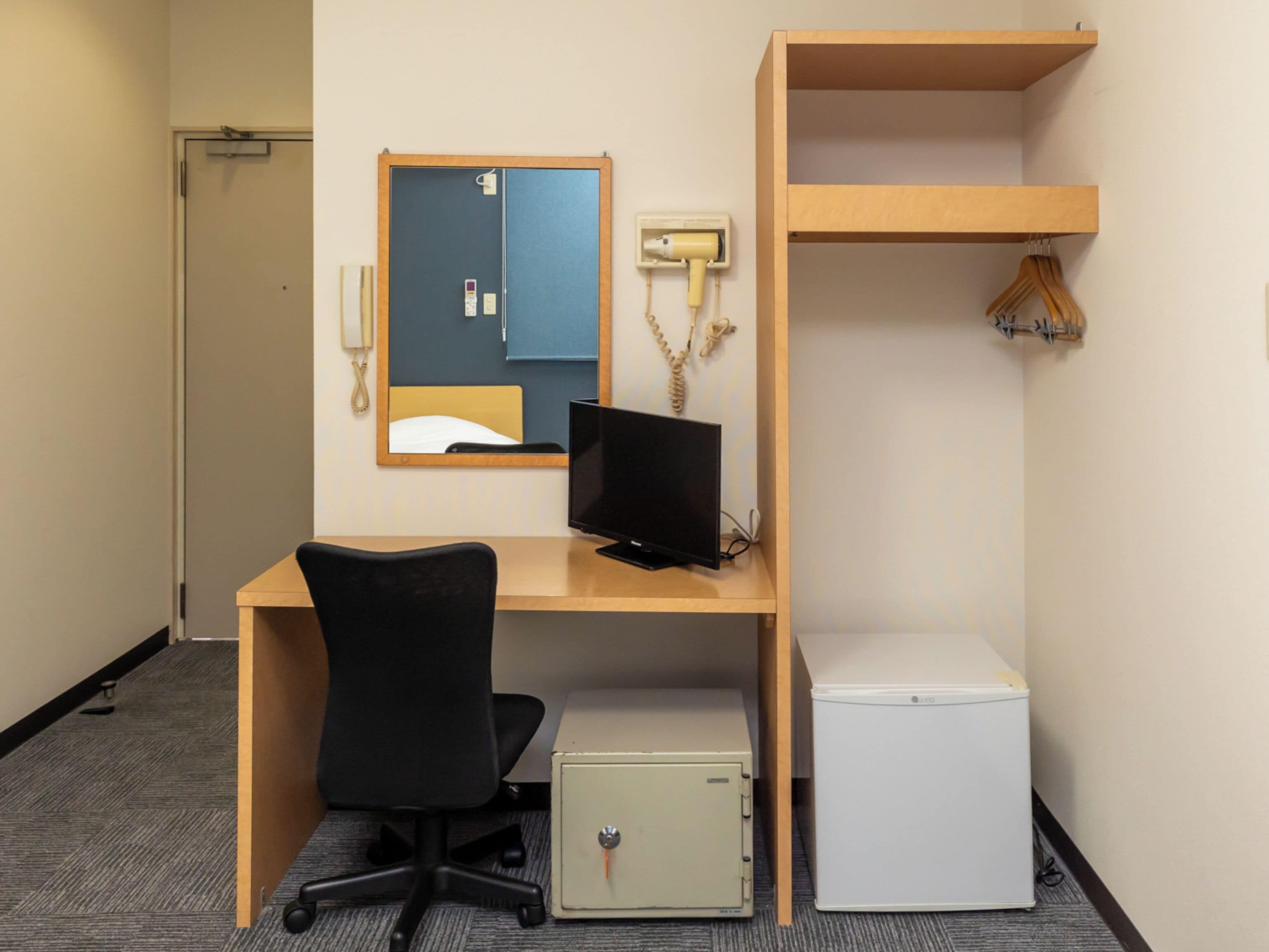 A hotel room with a desk, a computer, a chair, a small refrigerator, a mirror, a telephone, and a coat rack.