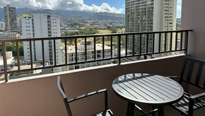 A balcony with a table and chairs, overlooking a cityscape with buildings and mountains in the distance. - Royal Kuhio 1602 - Spacious one‑bedroom condominium w/ Stunning Mountain Views in the Heart of Waikiki! (Waikiki)