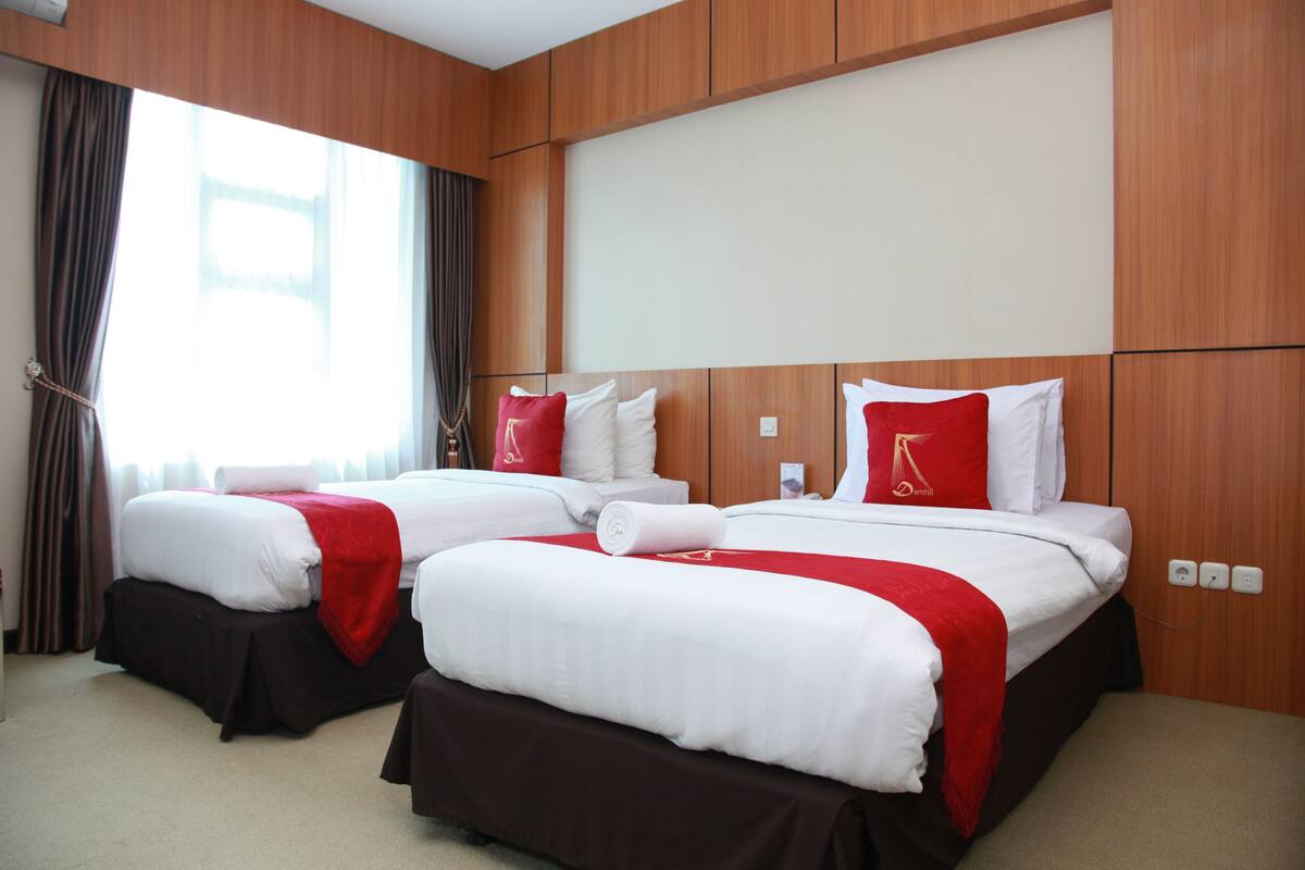 A hotel room with two beds, each with white bedding and red accents, wooden paneling on the walls, and a large window with curtains.