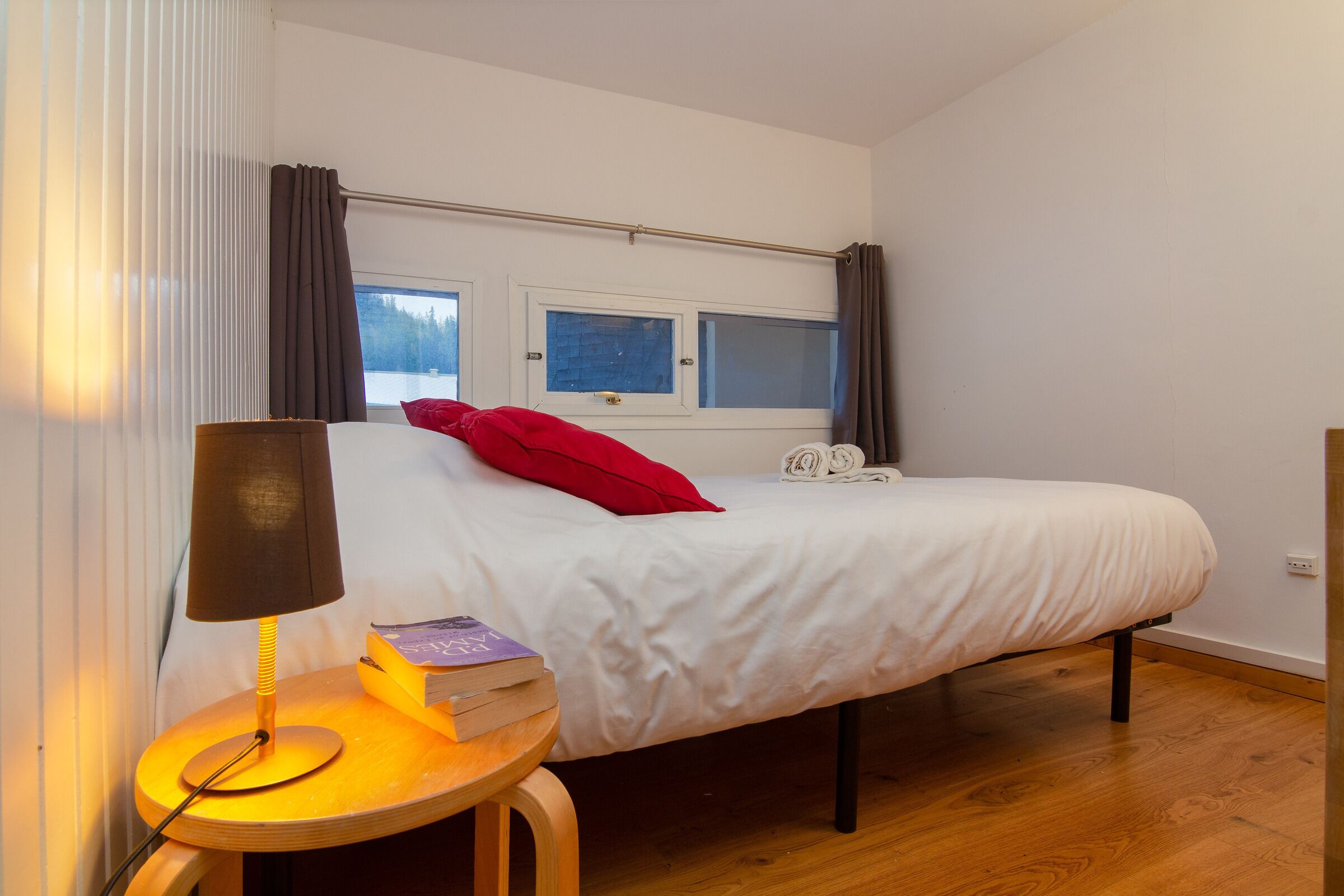 A bedroom with a bed, a bedside table with a lamp, a window, and a view of the outdoors.