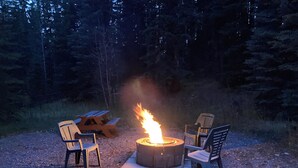 A campfire with two chairs and a picnic table in a forest setting. - Homesteader Cabin at Coliseum Mountain Resort (Nordegg)