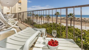 En balkon med hvide liggestole, et bord med en skål kirsebær og vinglas, samt udsigt over stranden og havet.