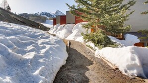 Un sentier enneigé mène à un bâtiment dont la porte est rouge, et qui est entouré de congères et d’un pin.