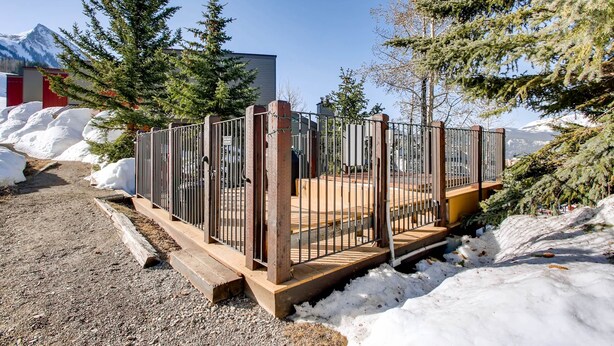 Une terrasse en bois avec des rampes en métal, entourée de neige et d’arbres, avec une montagne en arrière-plan.