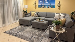 Ein Wohnzimmer mit einem grauen Ecksofa, einem kleinen runden Tisch mit Schiffsrad-Design, einem gemusterten Teppich und einem großen Fenster mit durchsichtigen Vorhängen.