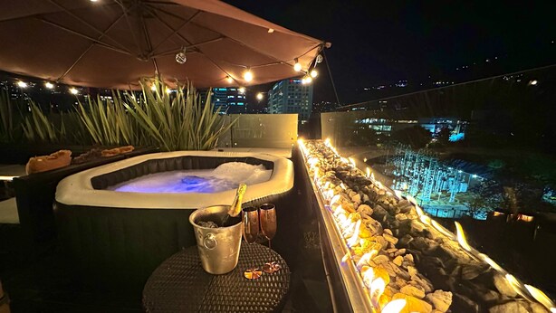 A hot tub with a champagne bottle, a bucket, and two glasses on a table, surrounded by plants and a stone wall with a pool. - Loft + private terrace with jacuzzi, fireplaces, cinema, panoramic view (Bogotá)