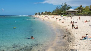 A beach with clear turquoise water, people swimming and sunbathing, and a building with a covered area in the background.