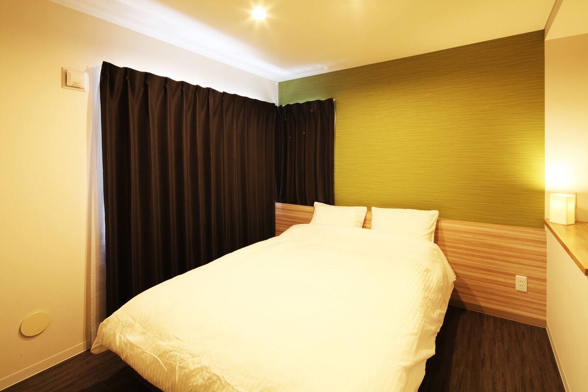 A neatly made bed in a hotel room with a wooden headboard, a large window with curtains, and a wall-mounted light.