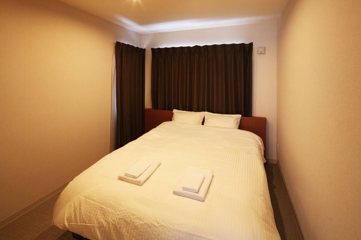 A hotel room with a neatly made bed, dark curtains, and a small bedside table.
