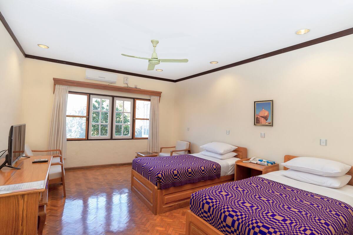 Une chambre d’hôtel avec deux lits, un bureau, une chaise, une télévision, un ventilateur de plafond et une fenêtre avec des rideaux.