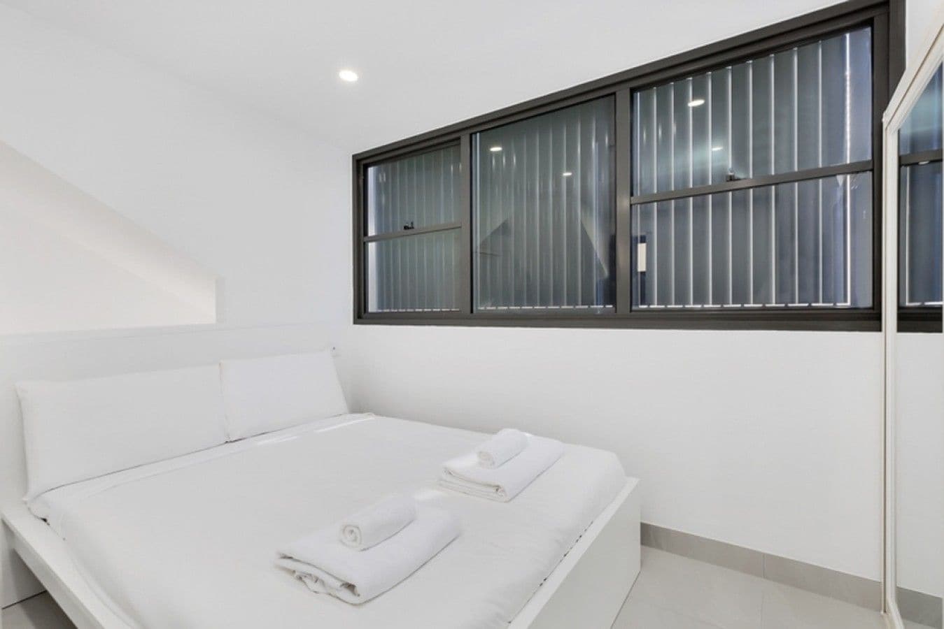 A modern hotel room with a single bed, two pillows, a window with blinds, and a white wall.