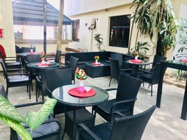 An outdoor seating area with round tables and black wicker chairs, featuring potted plants and flower arrangements.
