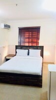 A neatly made bed in a room with a wooden headboard, a window with blinds, a bedside table, and an air conditioning unit on the wall.