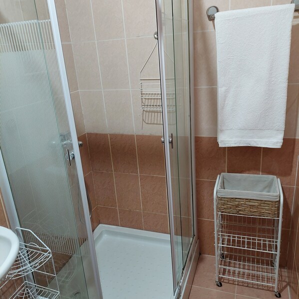 A shower area with a glass door, a white towel hanging on the wall, and a white trash bin.