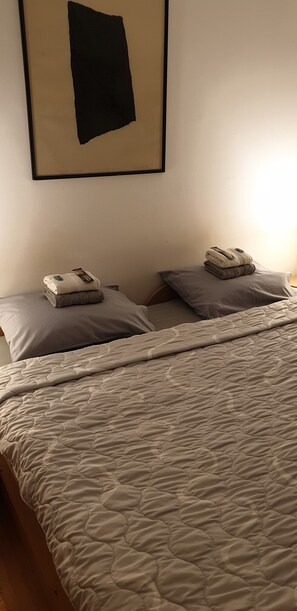 A neatly made bed with a quilted cover, two pillows, and a folded blanket on a wooden floor.