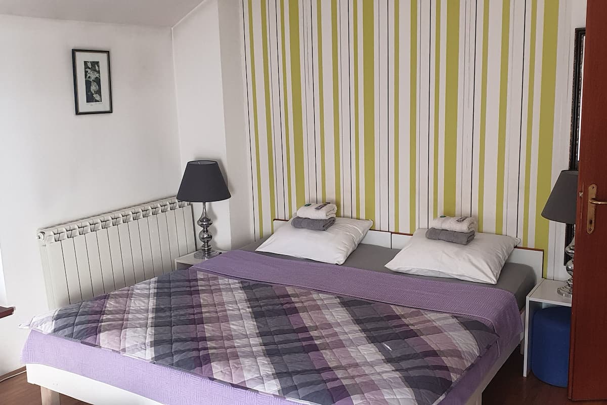 A bedroom with a bed, a nightstand, a lamp, a window with blinds, a door, and a striped wall.