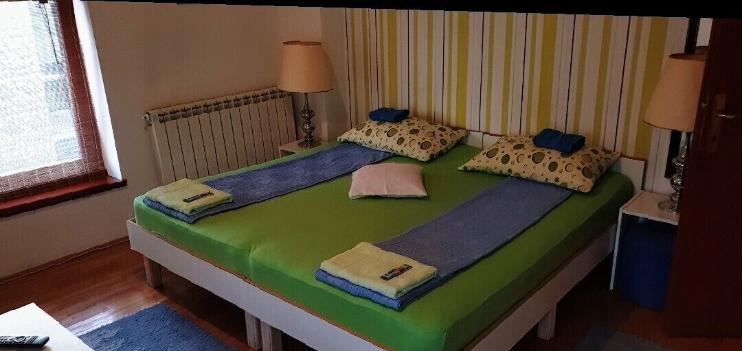 A single-bed room with a green bedspread, two pillows, a bedside lamp, and a window with blinds.