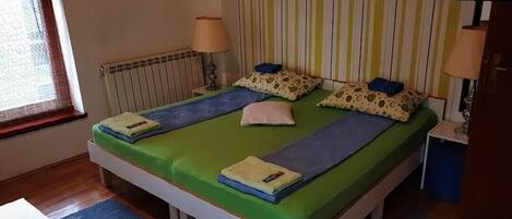 A single-bed room with a green bedspread, two pillows, a bedside lamp, and a window with blinds.
