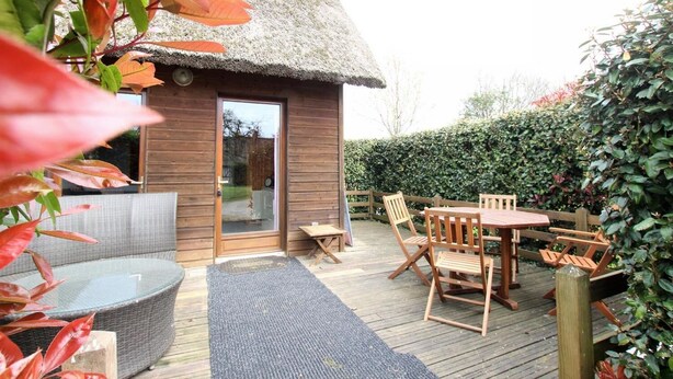 Un chalet au toit de chaume, doté d’une terrasse en bois, de meubles en osier et d’une porte en verre.