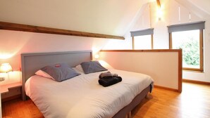 Une chambre avec un grand lit, deux tables de chevet, une commode, une poutre en bois au plafond et une fenêtre donnant sur un paysage verdoyant.