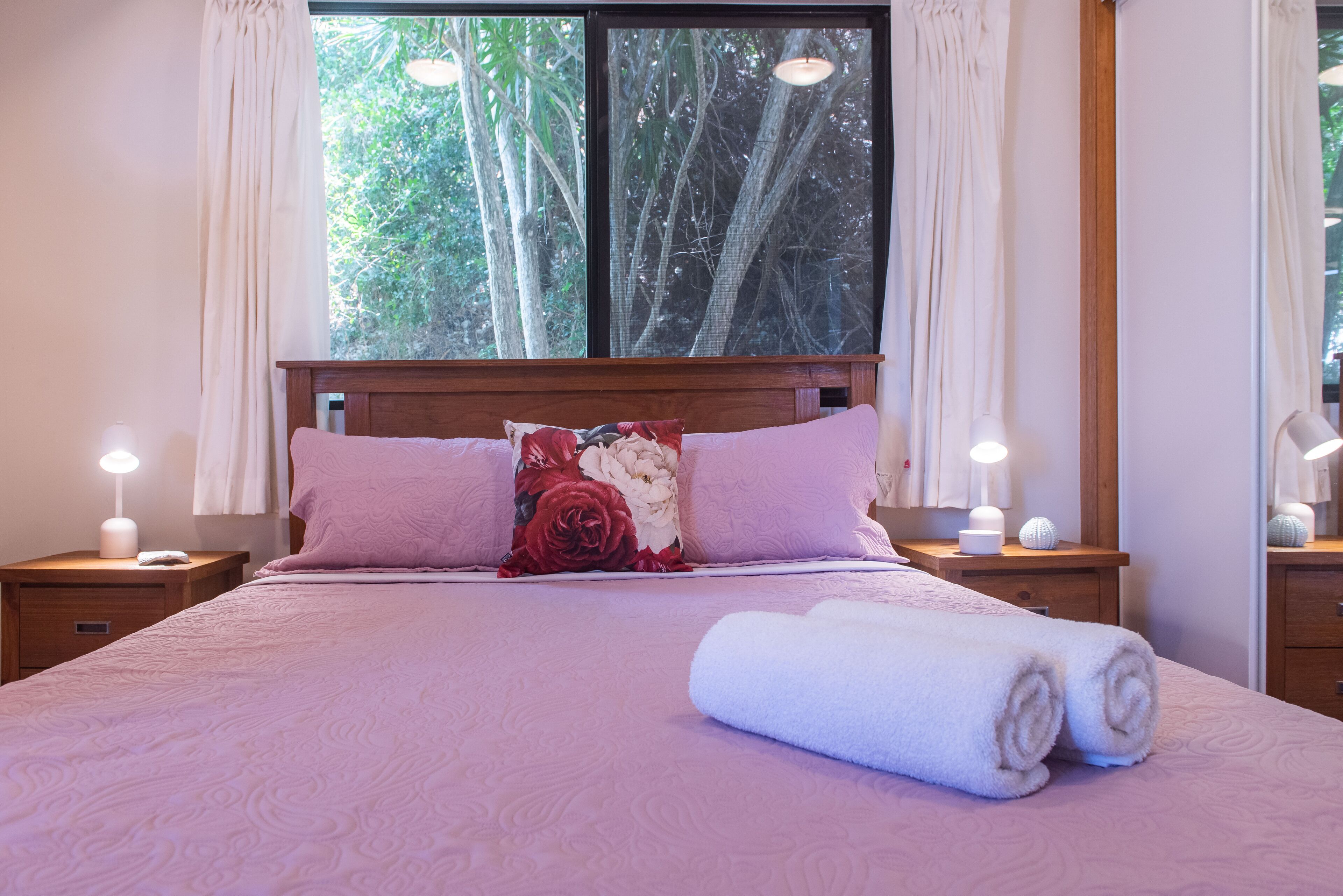 Een net opgemaakt bed met roze beddengoed en een bloemenkussen, twee nachtlampjes en uitzicht op bomen door een groot raam.